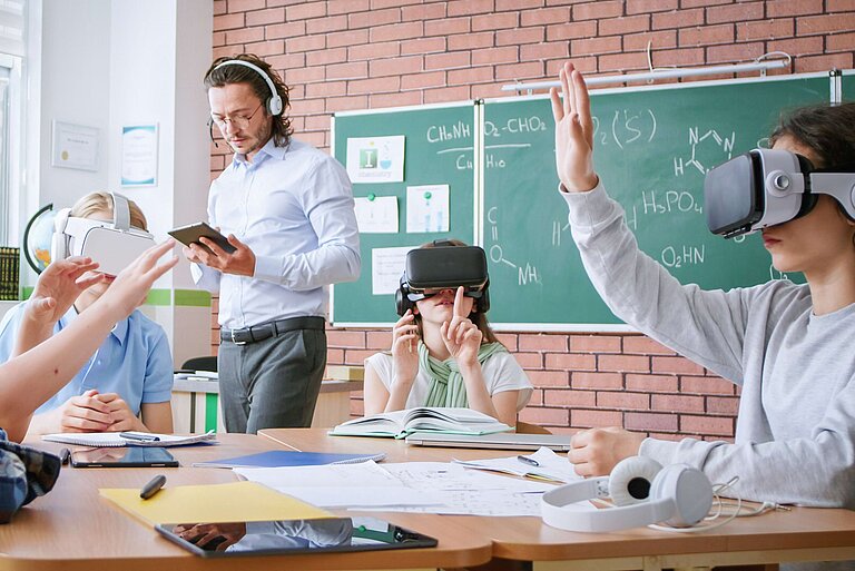 Sechs Schüler:innen sitzen mit VR-Brillen an einem Tisch in einem Klassenzimmer mit grünem Chemie-Tafelbild. Eine Person hebt die Hand, während eine Lehrkraft im Hintergrund mit einem Tablet interagiert. Auf dem Tisch liegen Notizbücher und Stifte.