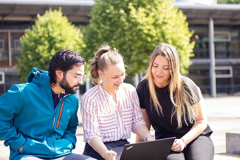 Drei junge Menschen sitzen draußen auf einer Bank oder Mauer vor einem modernen Gebäude mit großen Fenstern. Sie schauen gemeinsam auf einen Laptop und wirken konzentriert. Im Hintergrund grüne Bäume – Szene aus dem studentischen Alltag.