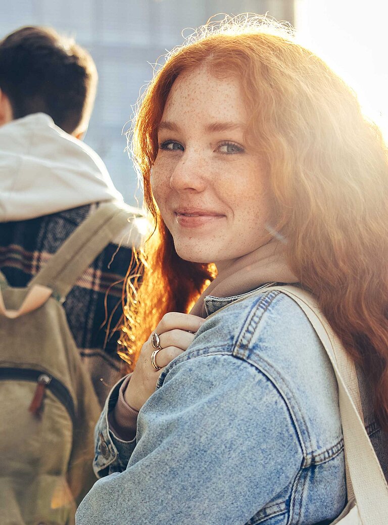 Fünf junge Menschen gehen gemeinsam durch ein urbanes Hochschulumfeld. Eine Person mit roten Haaren und Jeansjacke blickt lächelnd zurück zur Kamera, während die anderen mit Rucksäcken vorausgehen. Im Hintergrund moderne Gebäude und Sonnenlicht.