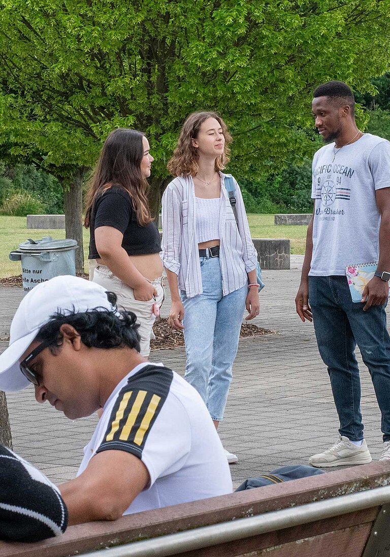 Junge Menschen in einem Parkähnlichen Außenbereich unter Bäumen; Gruppen unterhalten sich, einige sitzen auf Bänken, eine Person schreibt in ein Notizbuch; Rucksäcke deuten auf eine Campus- oder Schulumgebung hin.