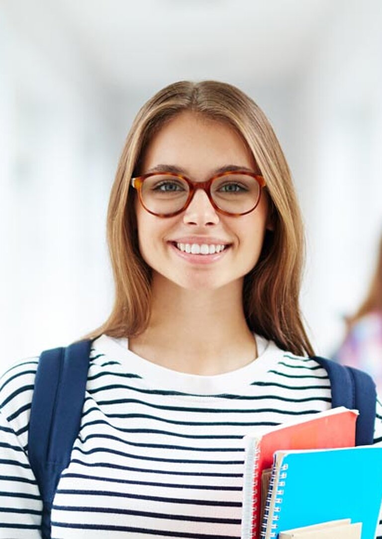 Junger Mensch mit Brille und gestreiftem Shirt steht lächelnd in einem hellen Flur mit großen Fenstern, hält Hefte und Ordner in der Hand. Im Hintergrund zwei weitere Personen im Gespräch – Szene aus der Hochschule Schmalkalden.